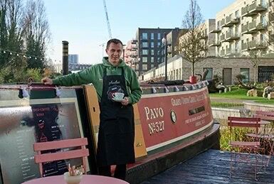 Man standing by a canal boat in Wembley with modern buildings in the background.