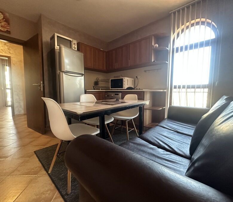 Interior of a 3-bedroom apartment with pool view, featuring a black leather sofa, dining area, and k.