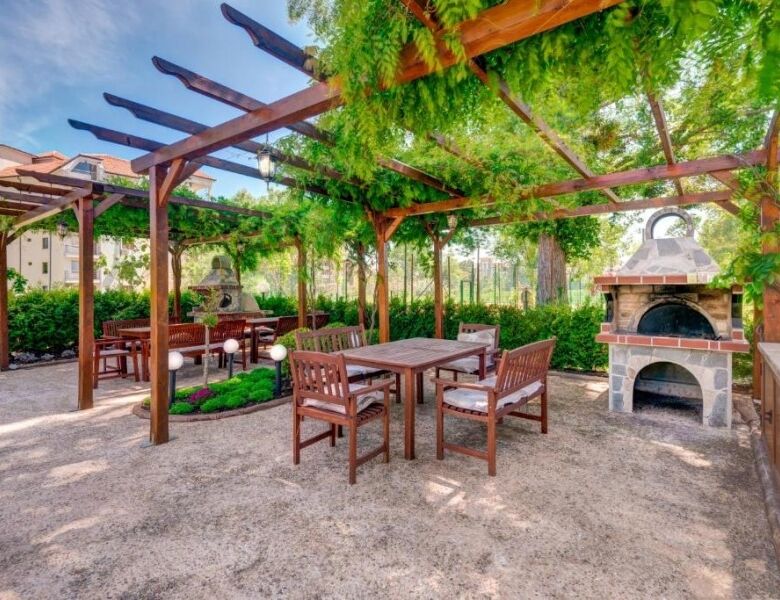 Outdoor dining space with wooden tables, chairs, and a stone pizza oven in a lush garden setting.