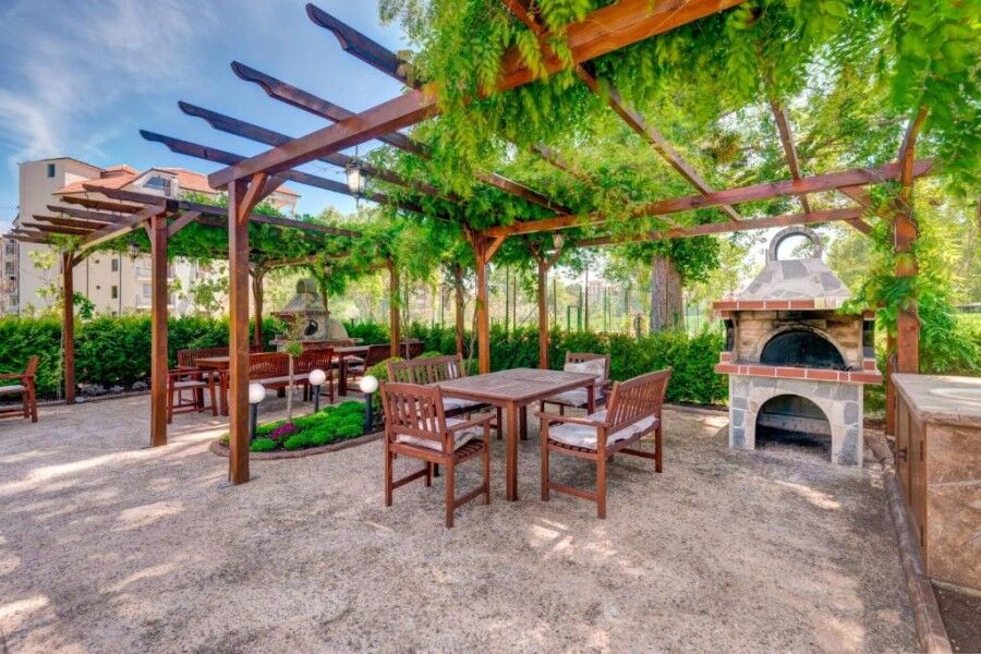 Outdoor dining space with wooden tables, chairs, and a stone pizza oven in a lush garden setting.