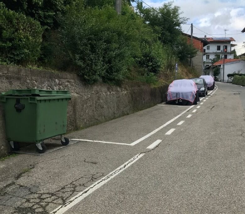 Parking area with cars and a green waste bin in a scenic Piedmont village setting.