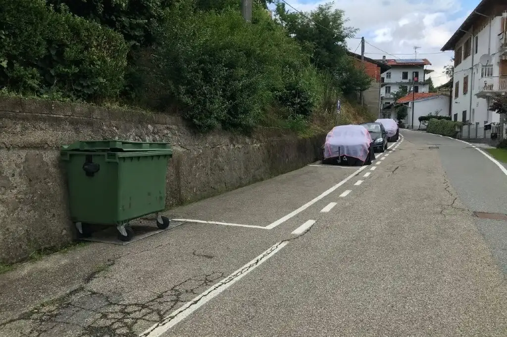 Parking area with cars and a green waste bin in a scenic Piedmont village setting.