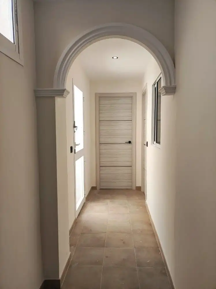 Arched doorway leading to a simple, bright hallway with white walls and beige flooring.