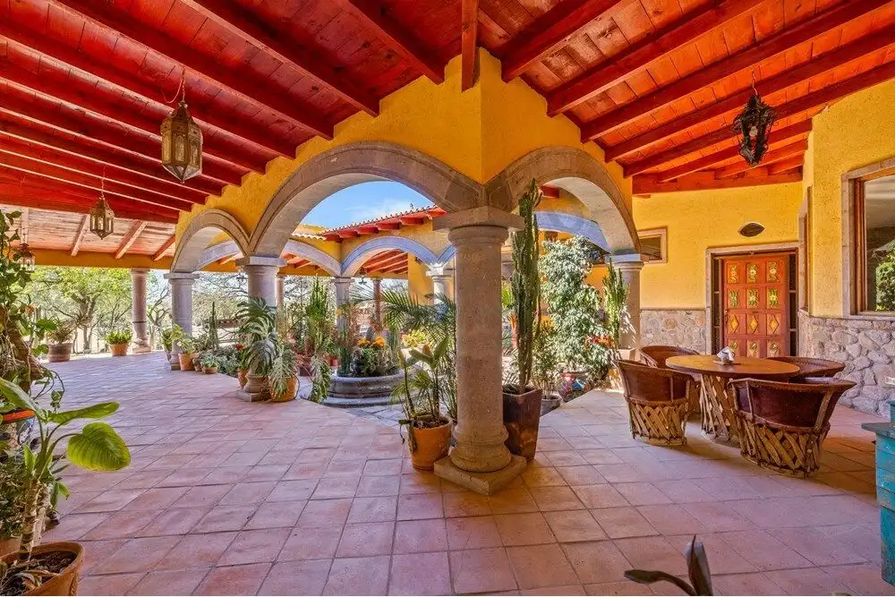 Spacious covered patio with arches, columns, and lush potted plants in a country estate setting.