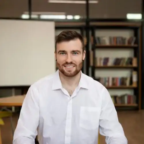Smiling man in a white shirt in a modern office setting with bookshelves.