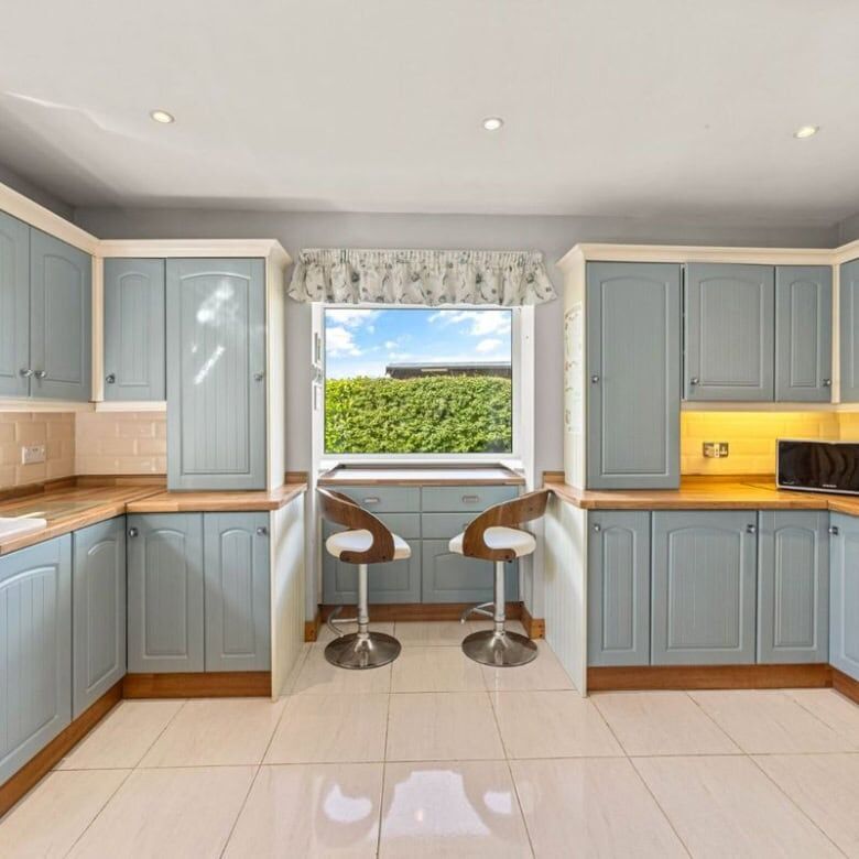 Bright blue kitchen with large window and two bar stools, modern appliances, and natural light.