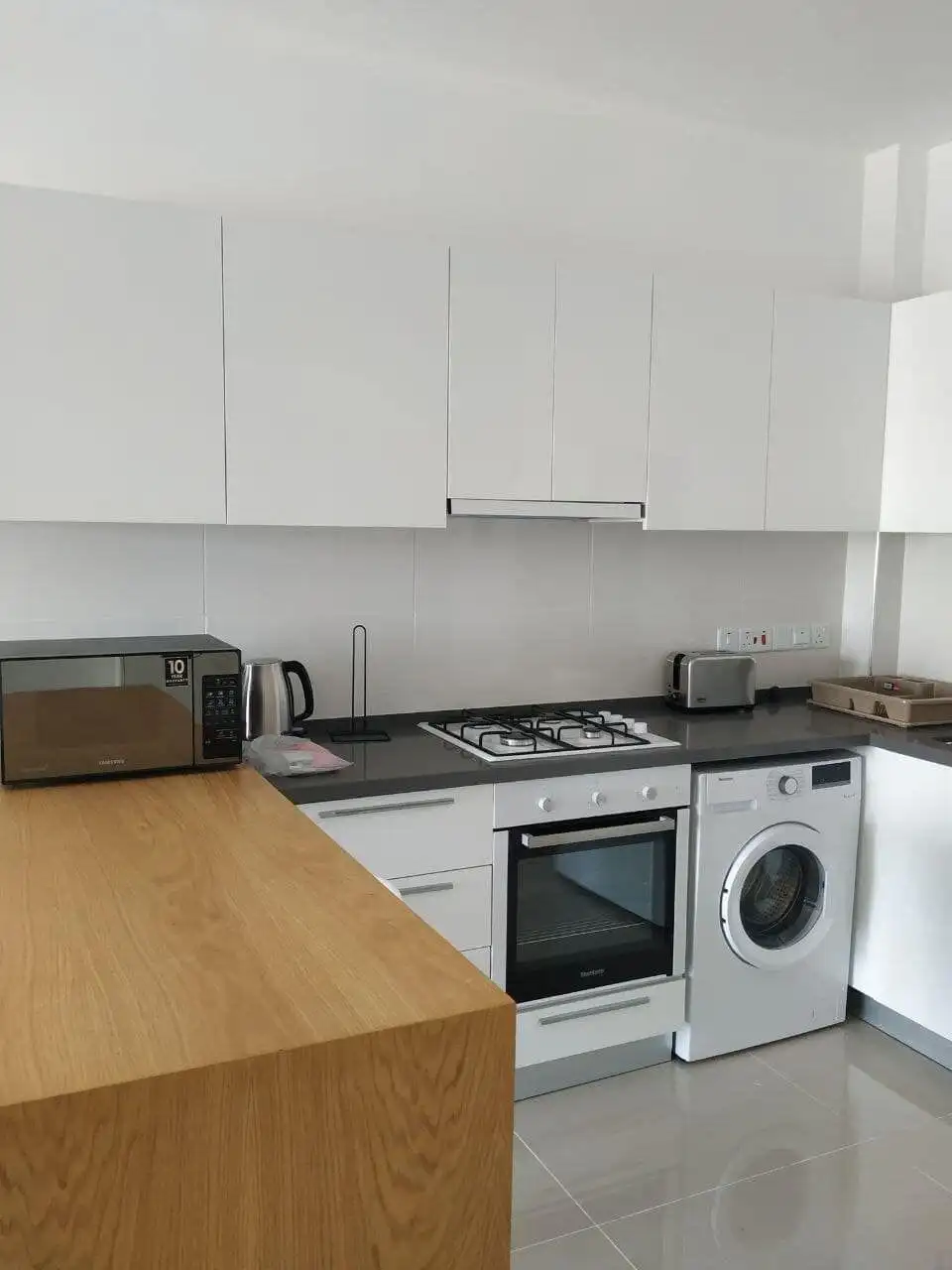 Modern kitchen with white cabinets, oven, washing machine, microwave, and countertop in Caeser’s Res.