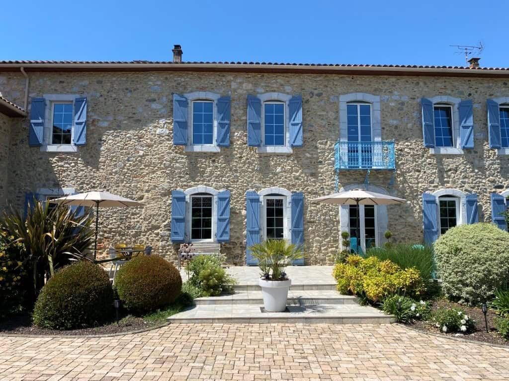 Built bed and breakfast with blue shutters, lush garden, and outdoor seating in Ausson, Haute Garonn.