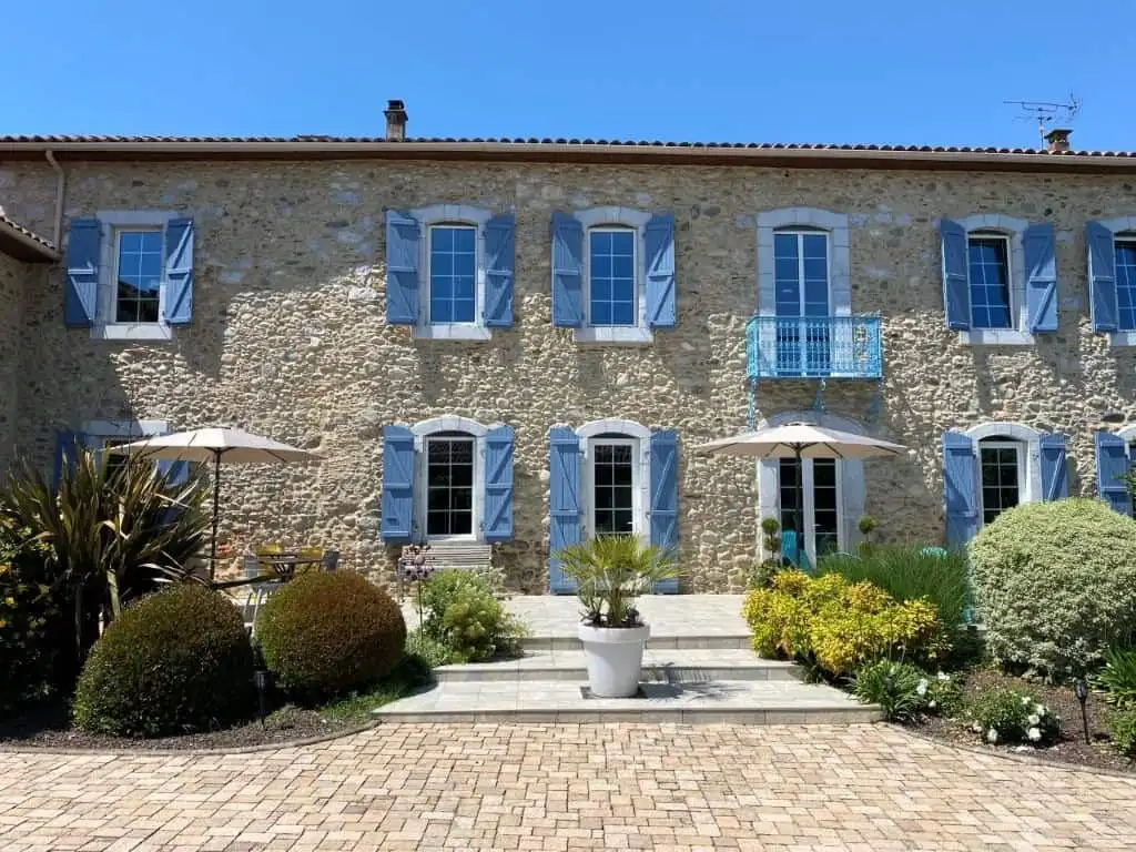 Built bed and breakfast with blue shutters, lush garden, and outdoor seating in Ausson, Haute Garonn.