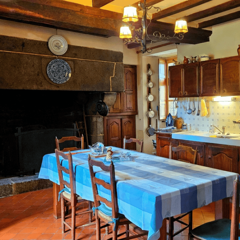 Cozy Normandy kitchen featuring a large fireplace, wooden cabinets, and a dining table with blue che.