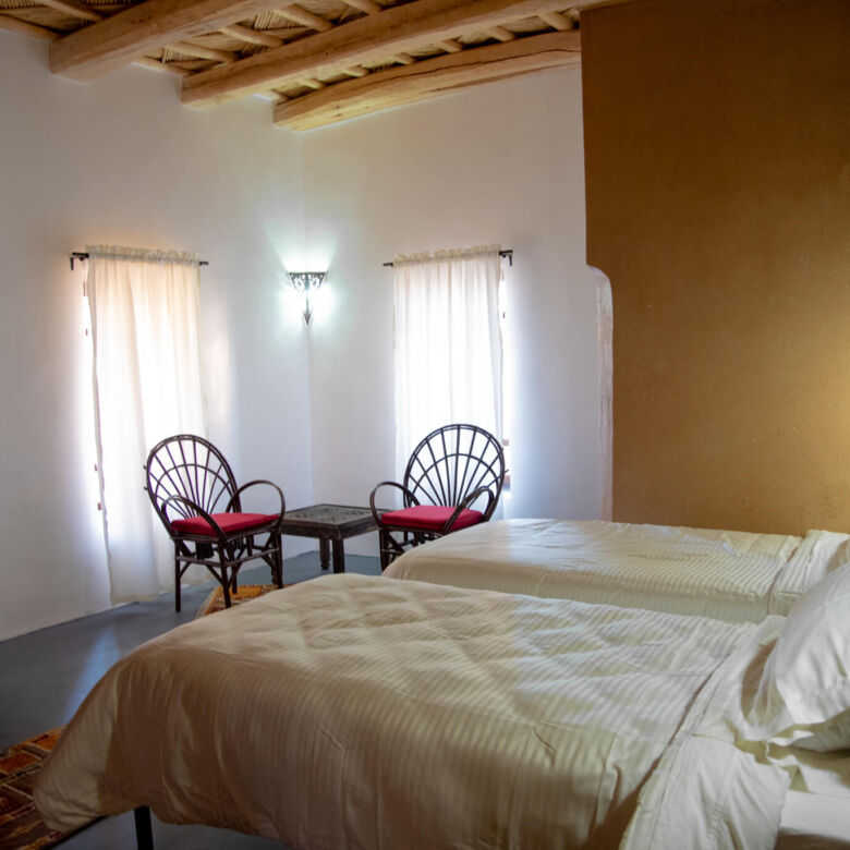 Cozy hotel bedroom with two beds, wooden ceiling, and natural light from windows.