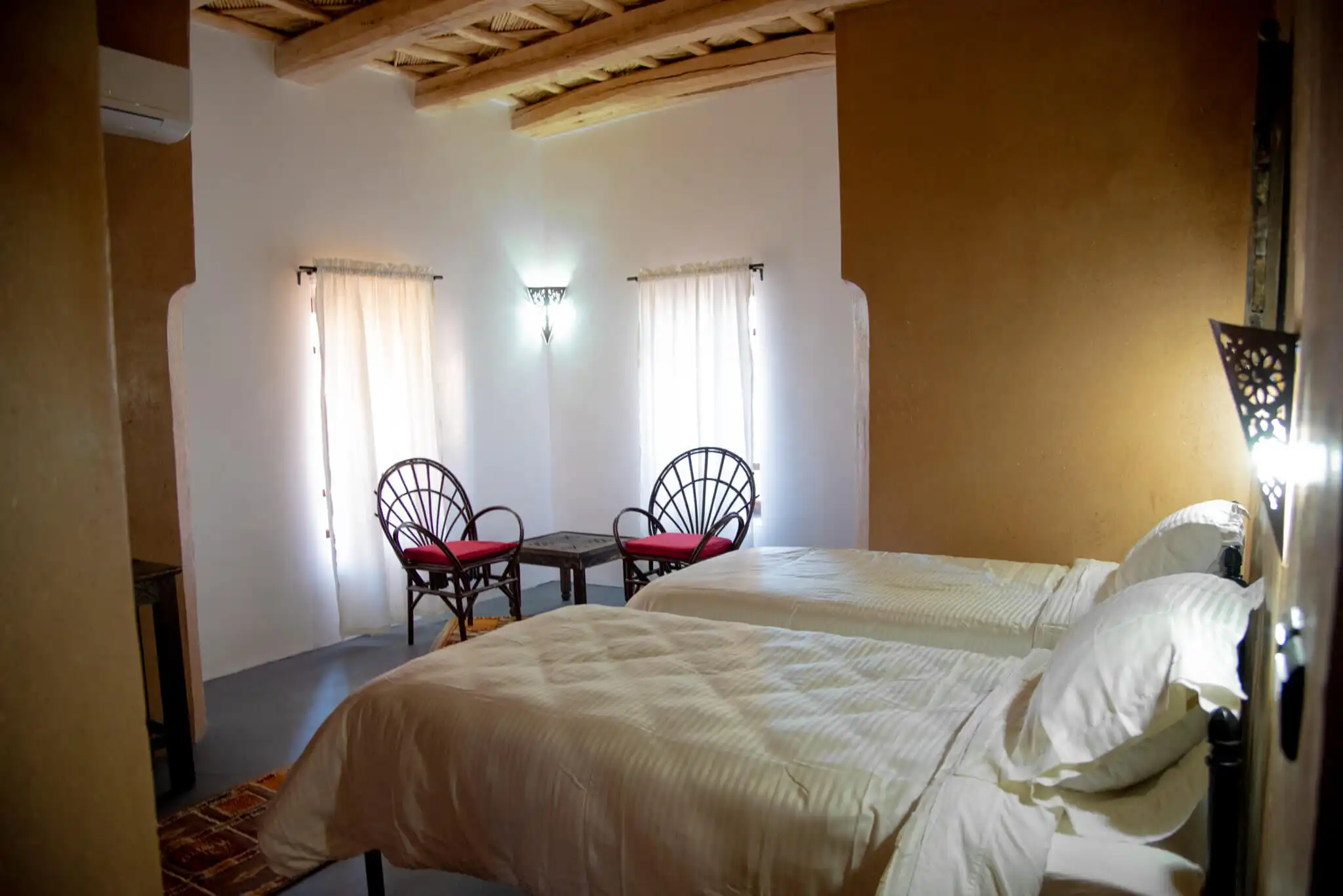 Cozy hotel bedroom with two beds, wooden ceiling, and natural light from windows.