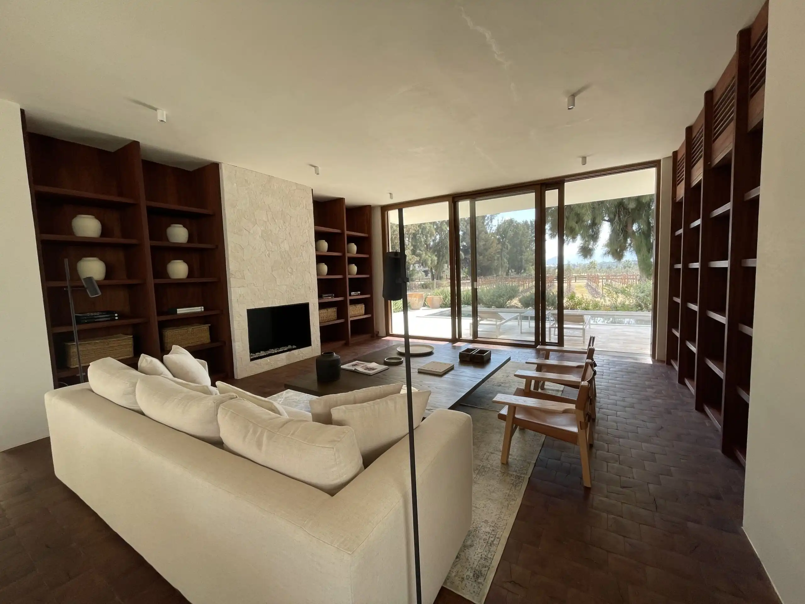 Modern living room with large windows and built-in wooden shelves. Spacious living area featuring a cozy sofa, wooden chairs, and a fireplace with ample natural light.