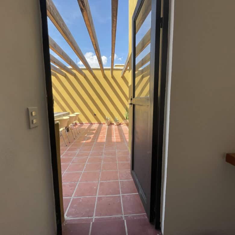 Balcony door opening to a sunny outdoor terrace with yellow and black striped railing.