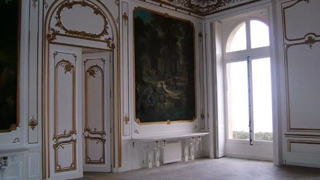 Luxurious château interior featuring ornate gold-trimmed walls, a large window, and classical furnis.