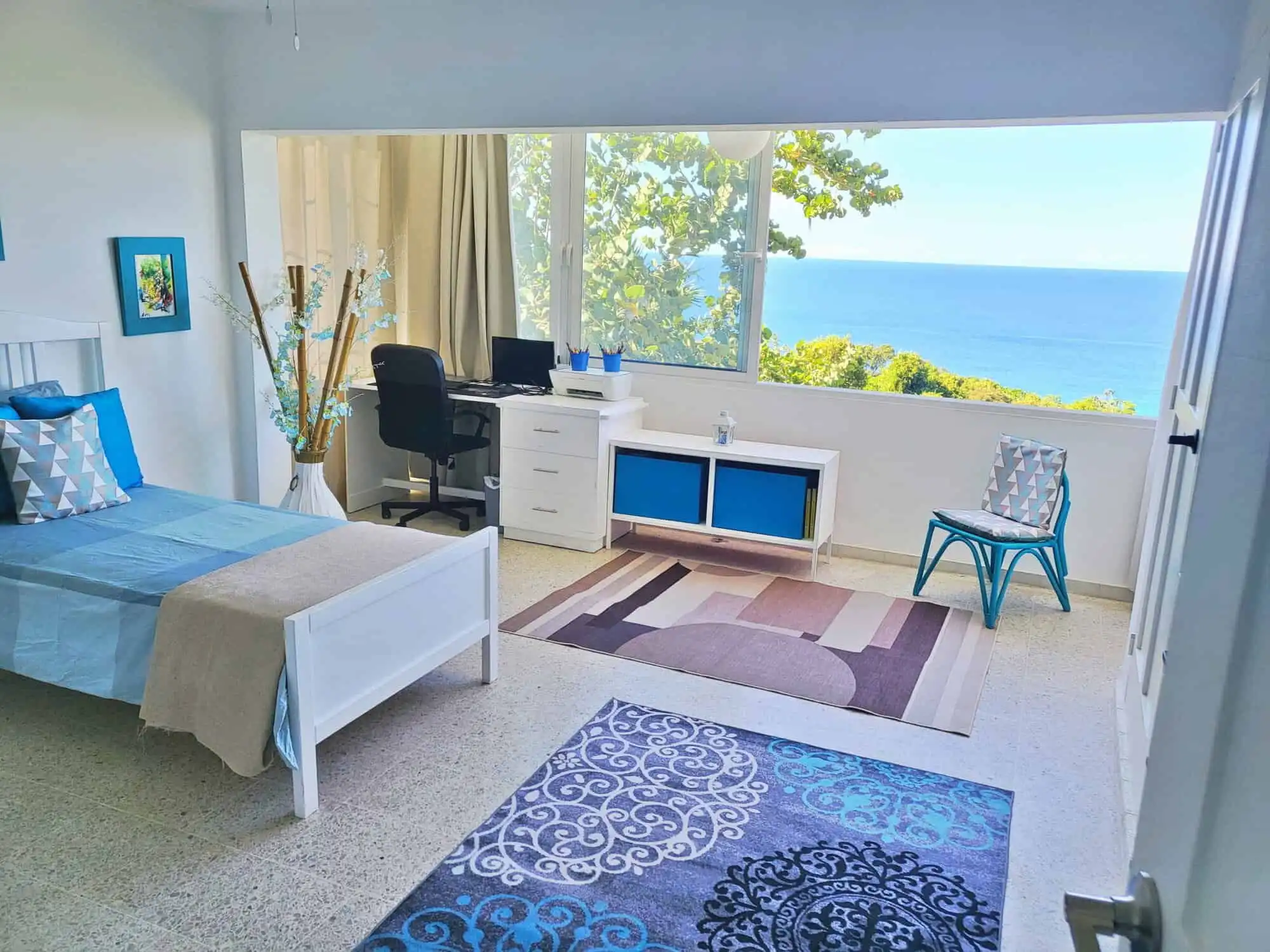 Bright ocean view room with large window overlooking the sea and lush greenery.