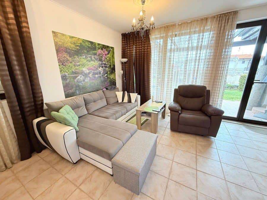 Modern living room with large windows and comfortable seating in a ground floor apartment. Bright living room with sofa, armchair, and scenic garden view through glass doors.