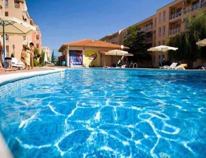 Pool with clear blue water at a sunny apartment complex with umbrellas and residents relaxing.