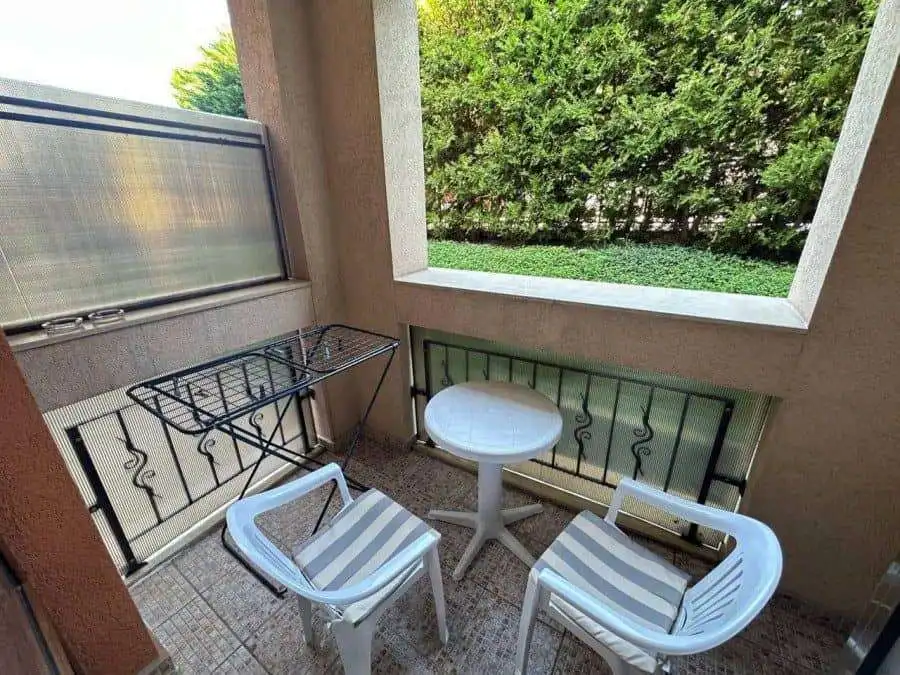 Balcony with two chairs, small table, and lush green background at Prestige Fort Beach.