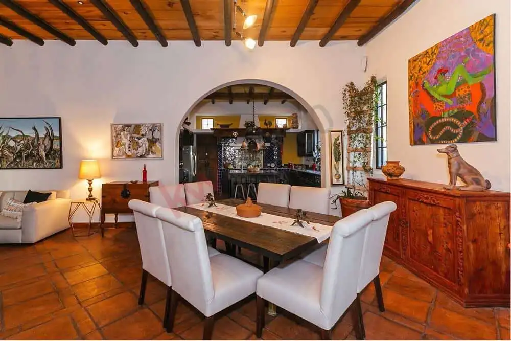 Elegant dining room with rustic wooden ceiling and vibrant artwork on walls. Spacious interior featuring a wooden dining table, white chairs, and colorful art pieces.