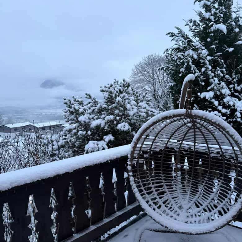 Cozy hanging chair on snow-covered balcony overlooking snowy landscape and distant mountains.