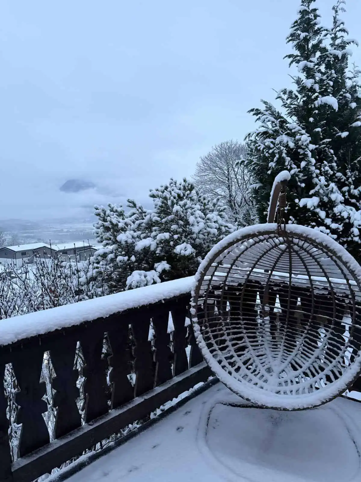 Cozy hanging chair on snow-covered balcony overlooking snowy landscape and distant mountains.