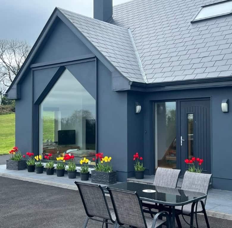 Contemporary blue house with large glass windows and colorful potted plants outside.