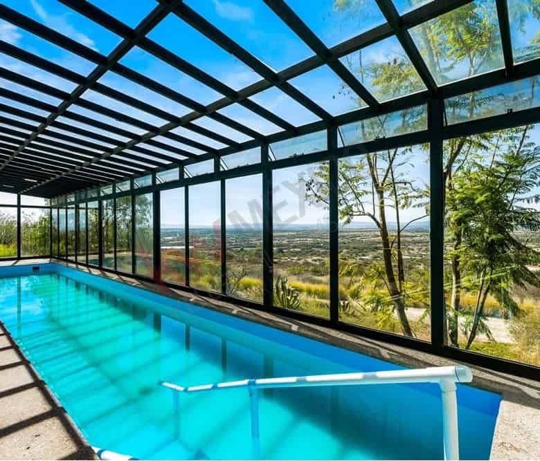 Indoor pool with glass walls overlooking lush landscape and clear blue sky.
