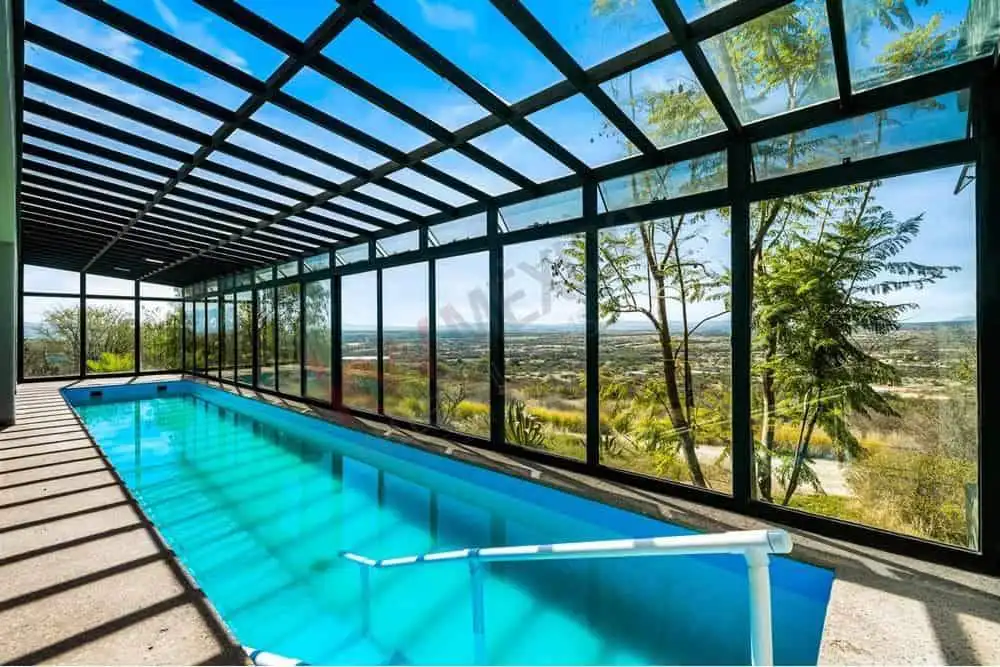 Indoor pool with glass walls overlooking lush landscape and clear blue sky.