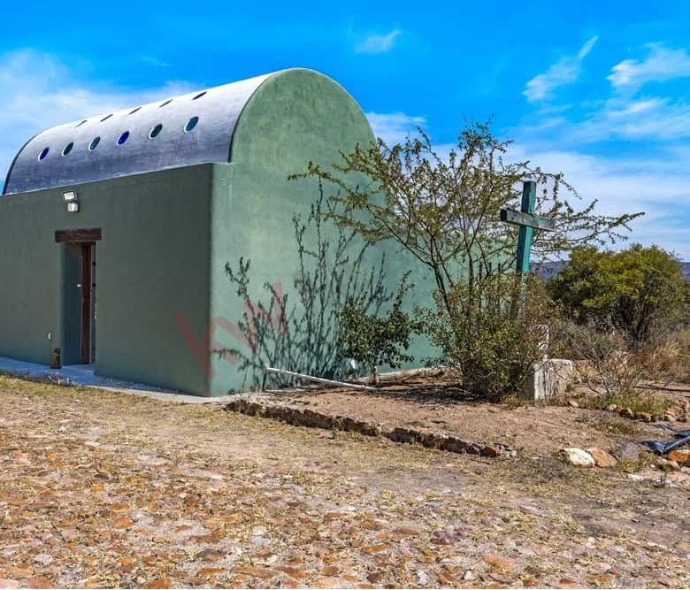 Small green house with a curved roof in a rural setting, surrounded by dry vegetation and a clear bl.