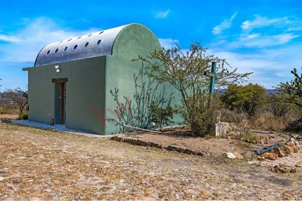 Small green house with a curved roof in a rural setting, surrounded by dry vegetation and a clear bl.