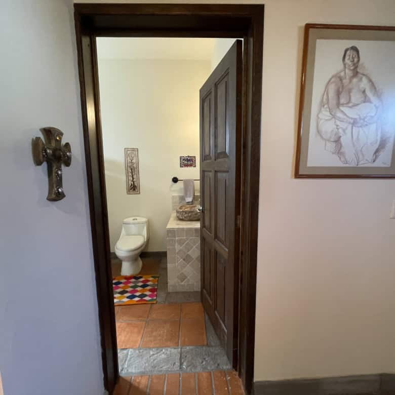 Home interior with bathroom visible from hallway, featuring a wooden door, artwork, and tiled floori.