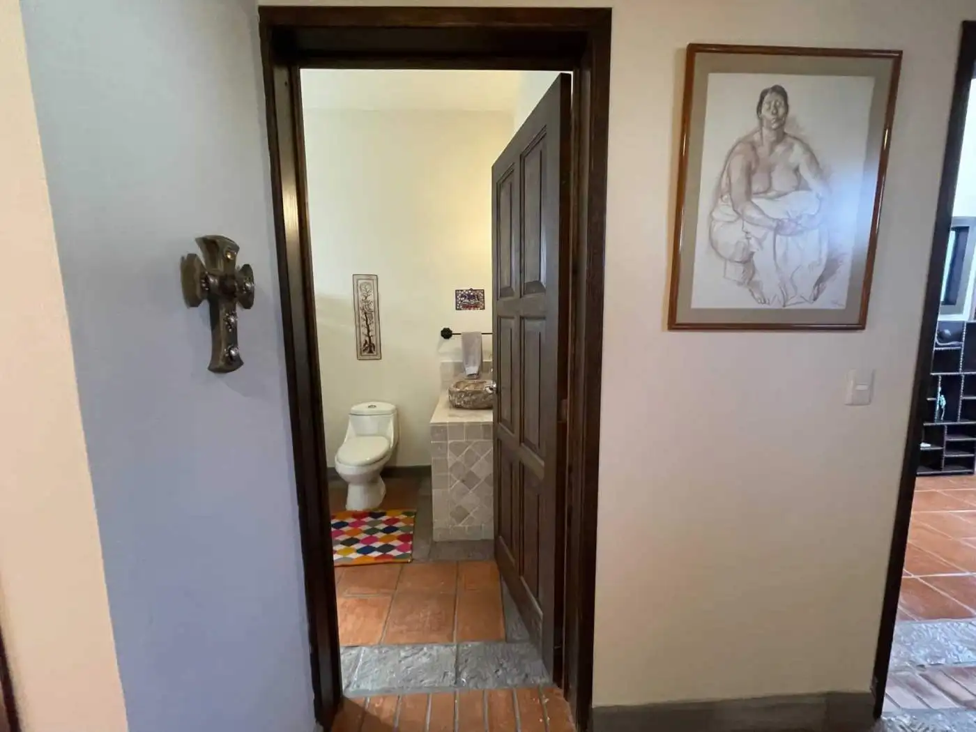 Home interior with bathroom visible from hallway, featuring a wooden door, artwork, and tiled floori.