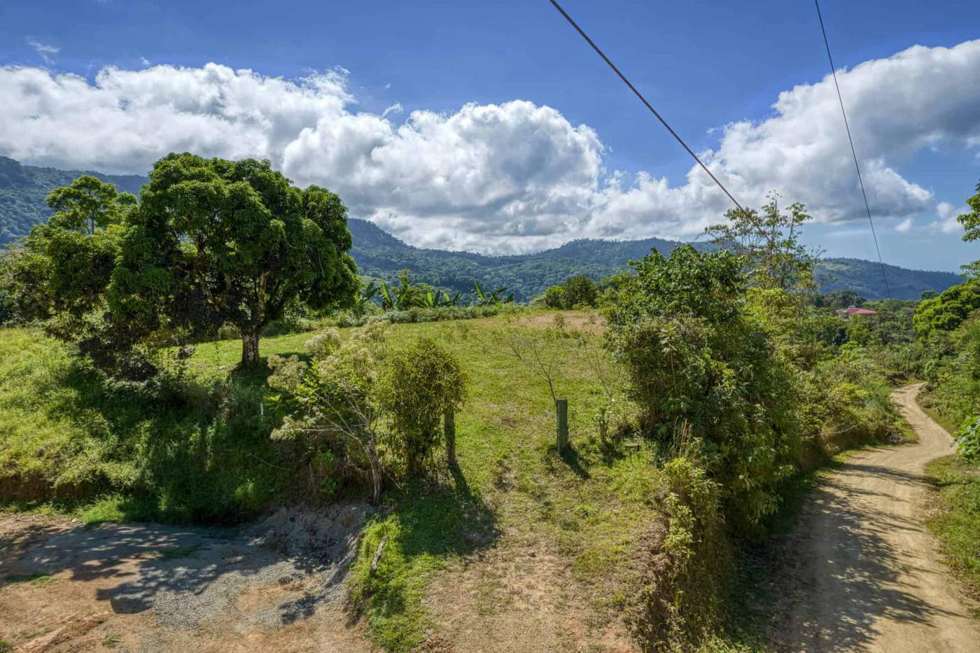 Rural landscape with trees, green hills, and a dirt path under a partly cloudy sky.