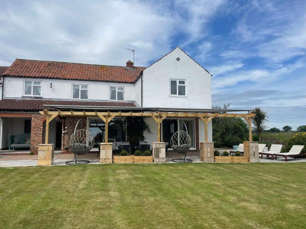 Modern countryside house with outdoor seating area and garden. Country house with outdoor patio, garden, and lounge chairs under a wooden pergola.