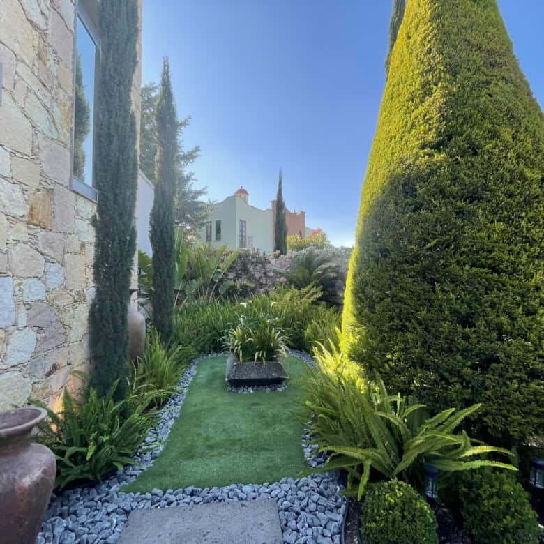 Elegant garden with lush greenery, trimmed bushes, and a stone house exterior under a clear blue sky.