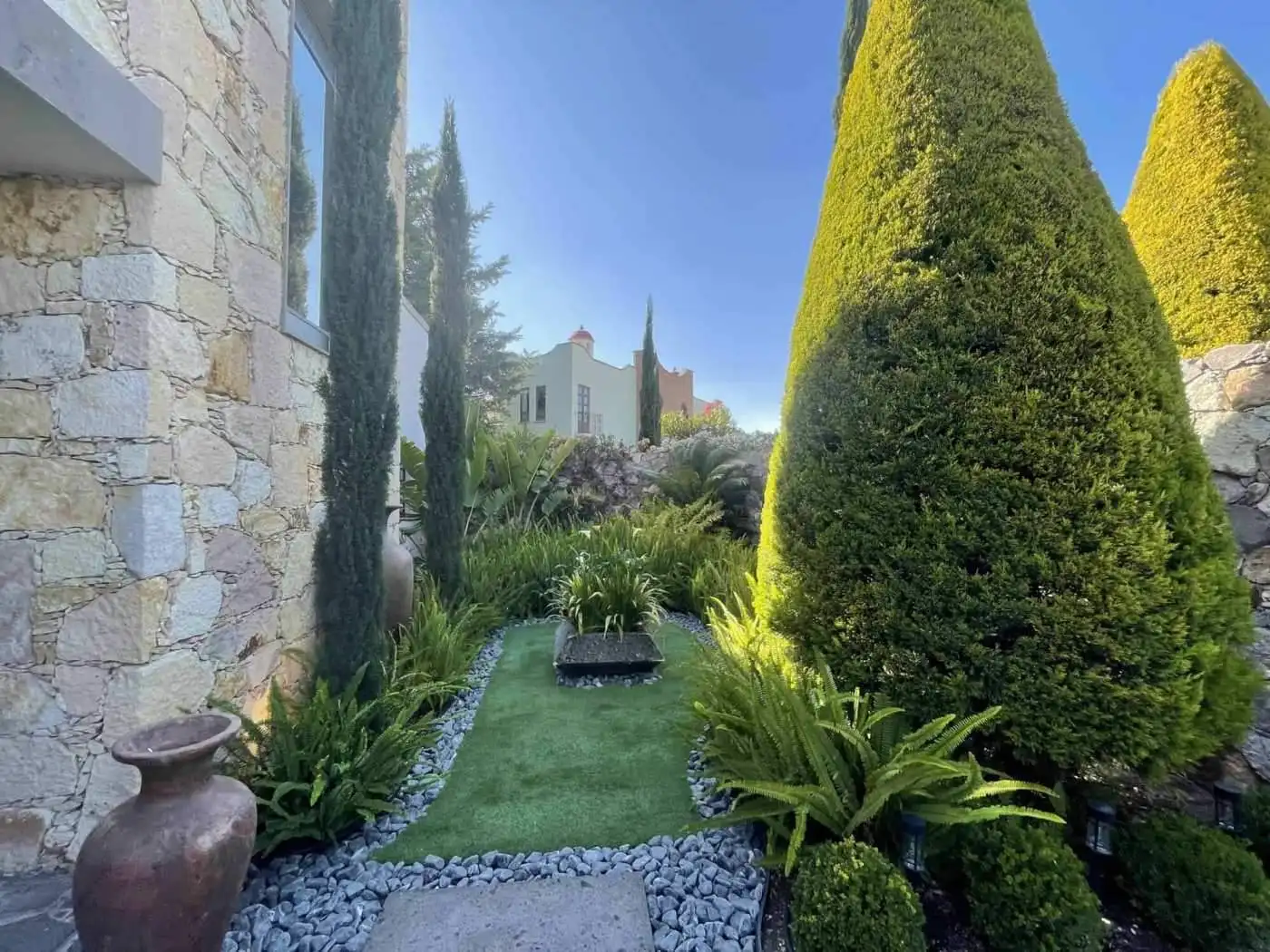 Elegant garden with lush greenery, trimmed bushes, and a stone house exterior under a clear blue sky.