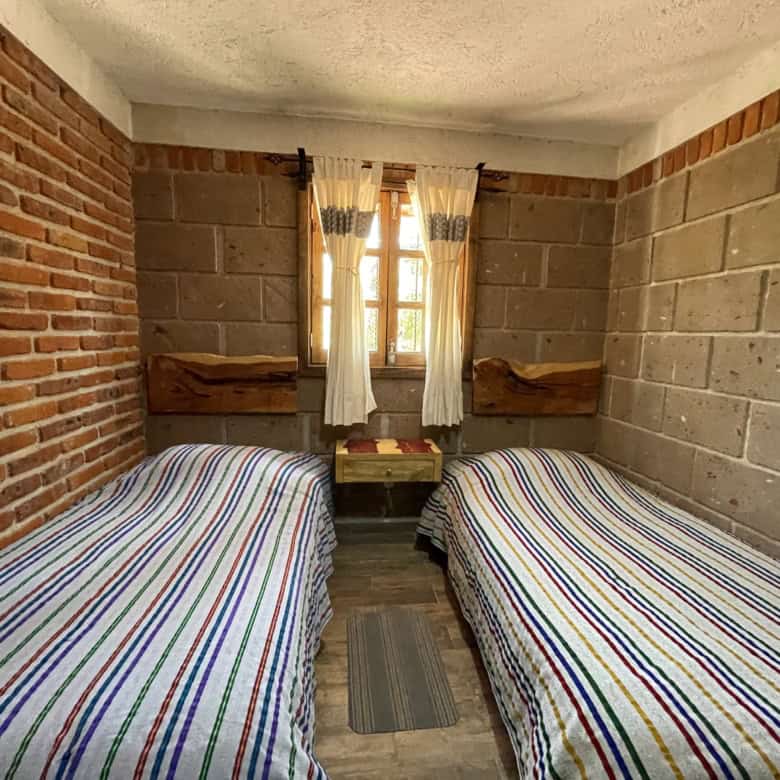 Small bedroom with two single beds, brick and concrete walls, and a window with curtains.