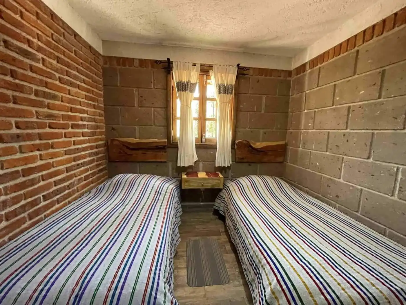 Small bedroom with two single beds, brick and concrete walls, and a window with curtains.