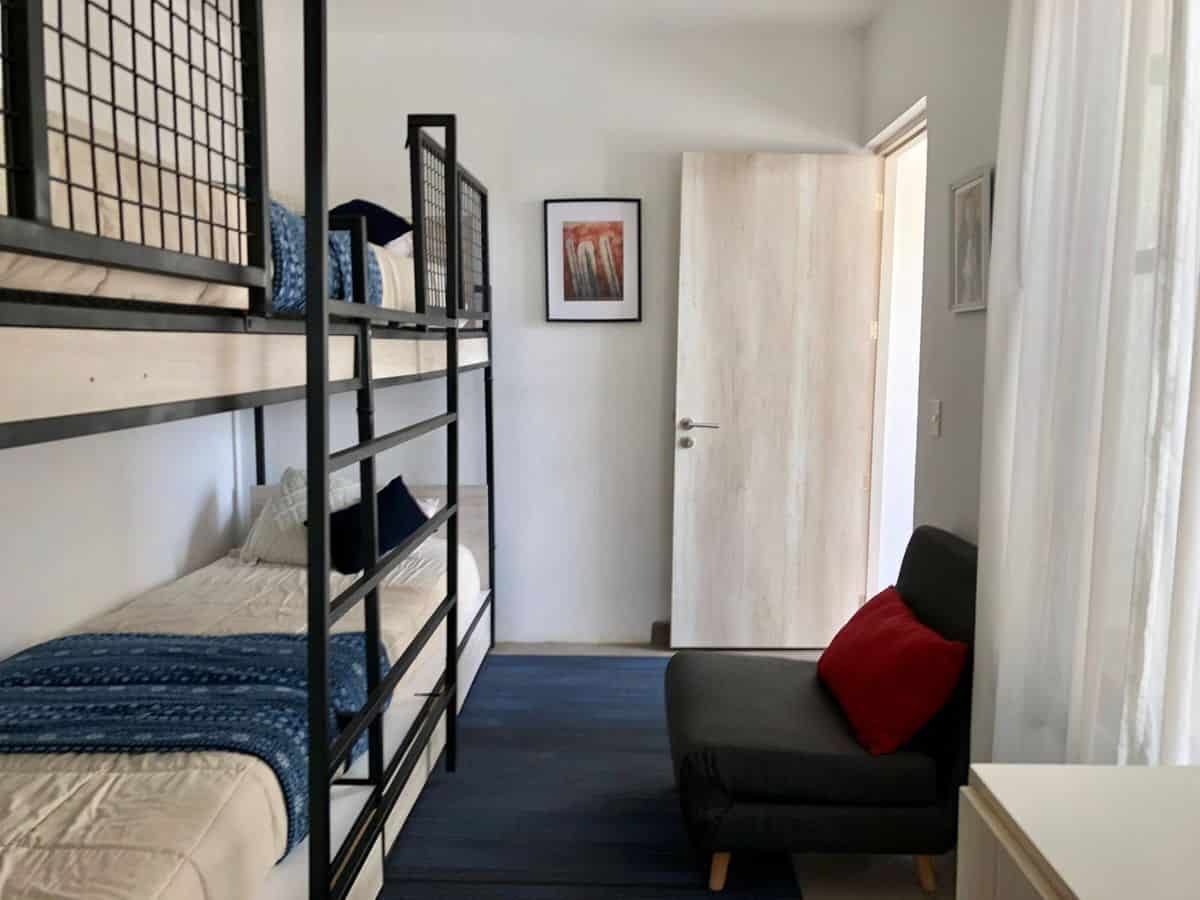 Bunk bed with blue bedding and a small black sofa with red cushion in a bright, minimalist bedroom.