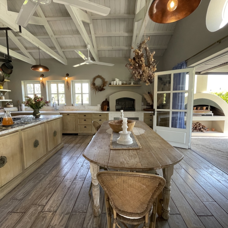 Rustic kitchen with wooden cabinets, open shelves, and a large dining table in a bright, airy space.