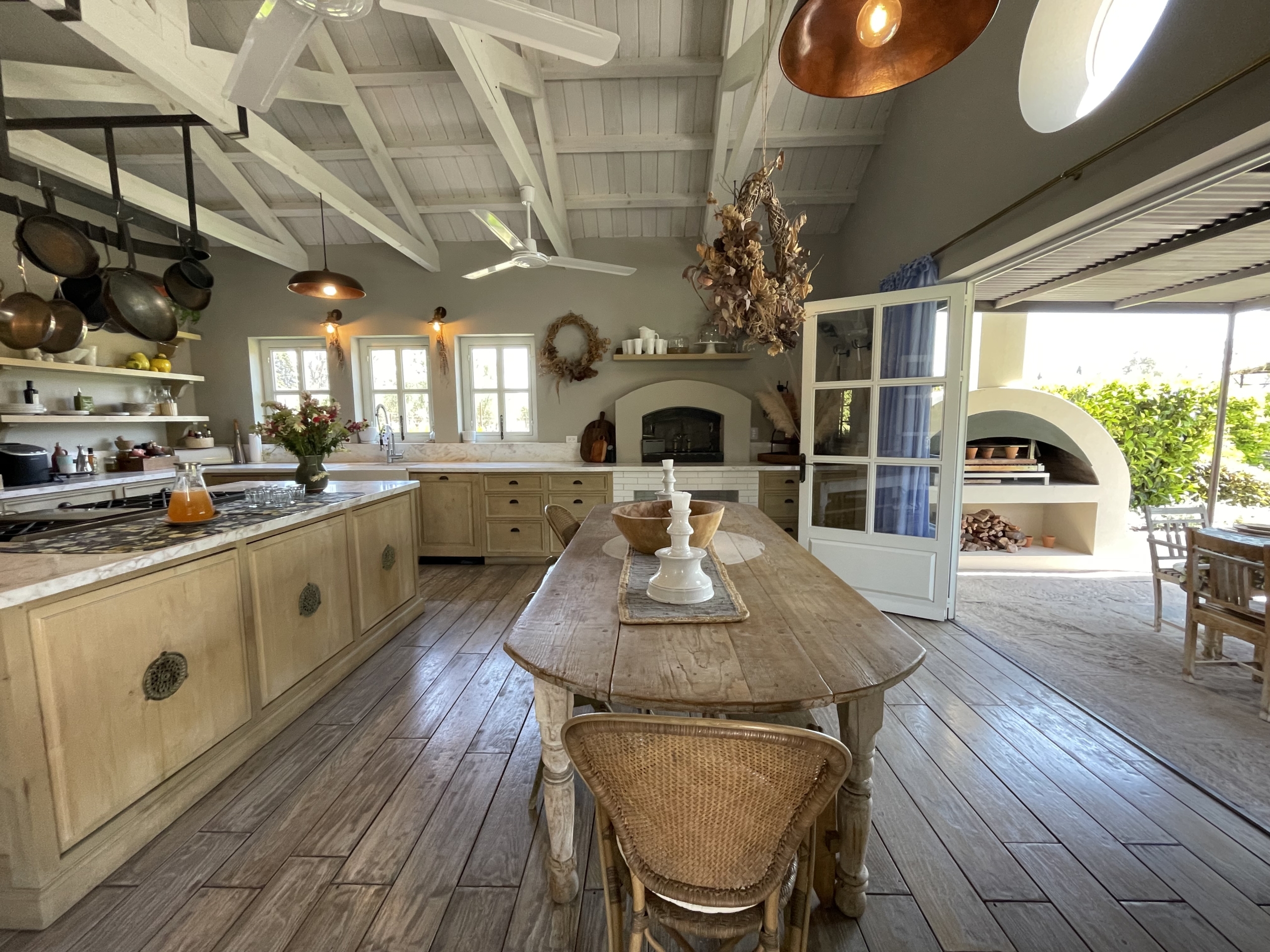Rustic kitchen with wooden cabinets, open shelves, and a large dining table in a bright, airy space.
