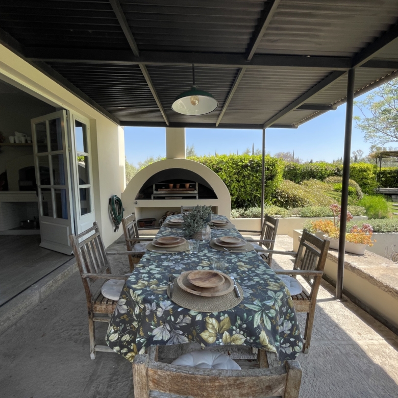 Outdoor dining space with table and chairs under a covered patio, overlooking a lush garden and sunn.