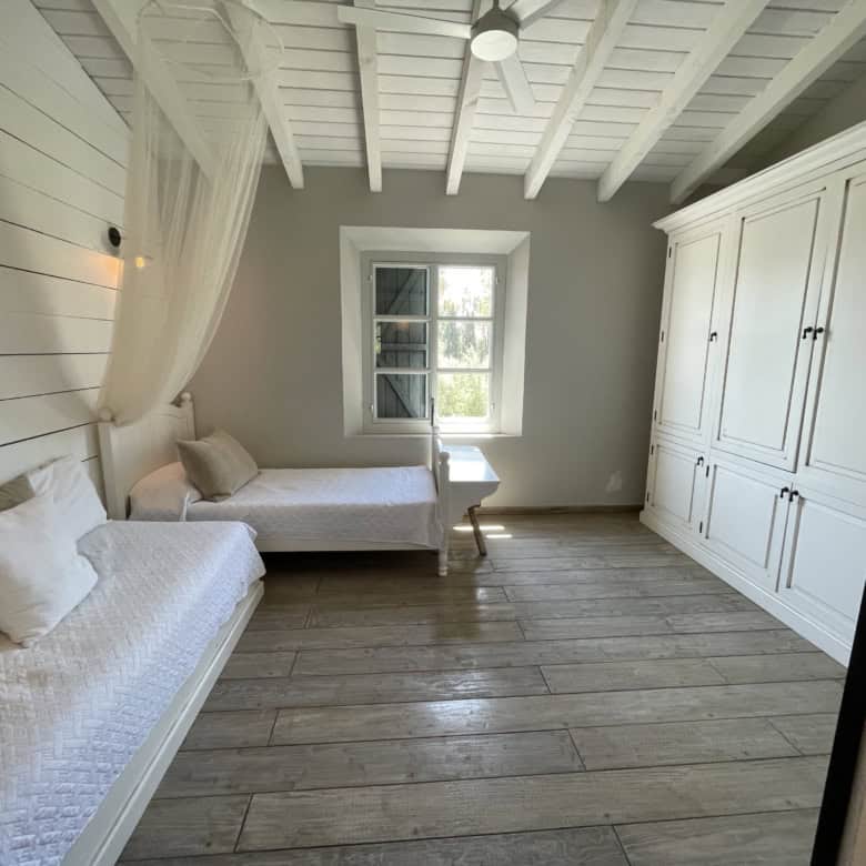 Bright coastal bedroom with white wooden walls and ceiling, featuring two single beds and a large wa.