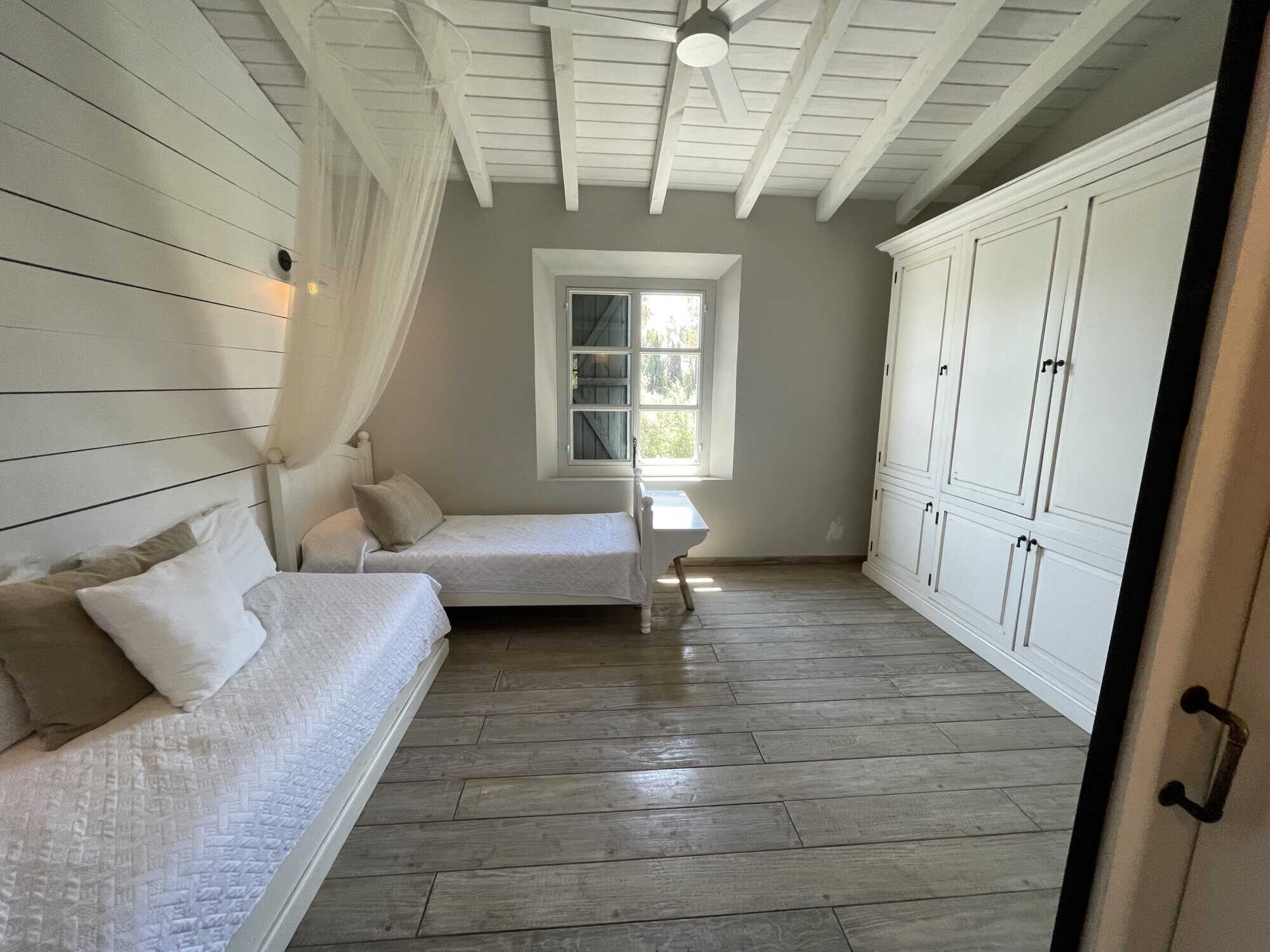 Bright coastal bedroom with white wooden walls and ceiling, featuring two single beds and a large wa.