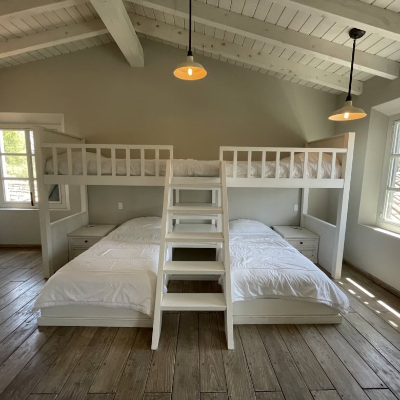 Attic bedroom with white bunk beds, large windows, and wooden flooring, offering a bright and inviti.