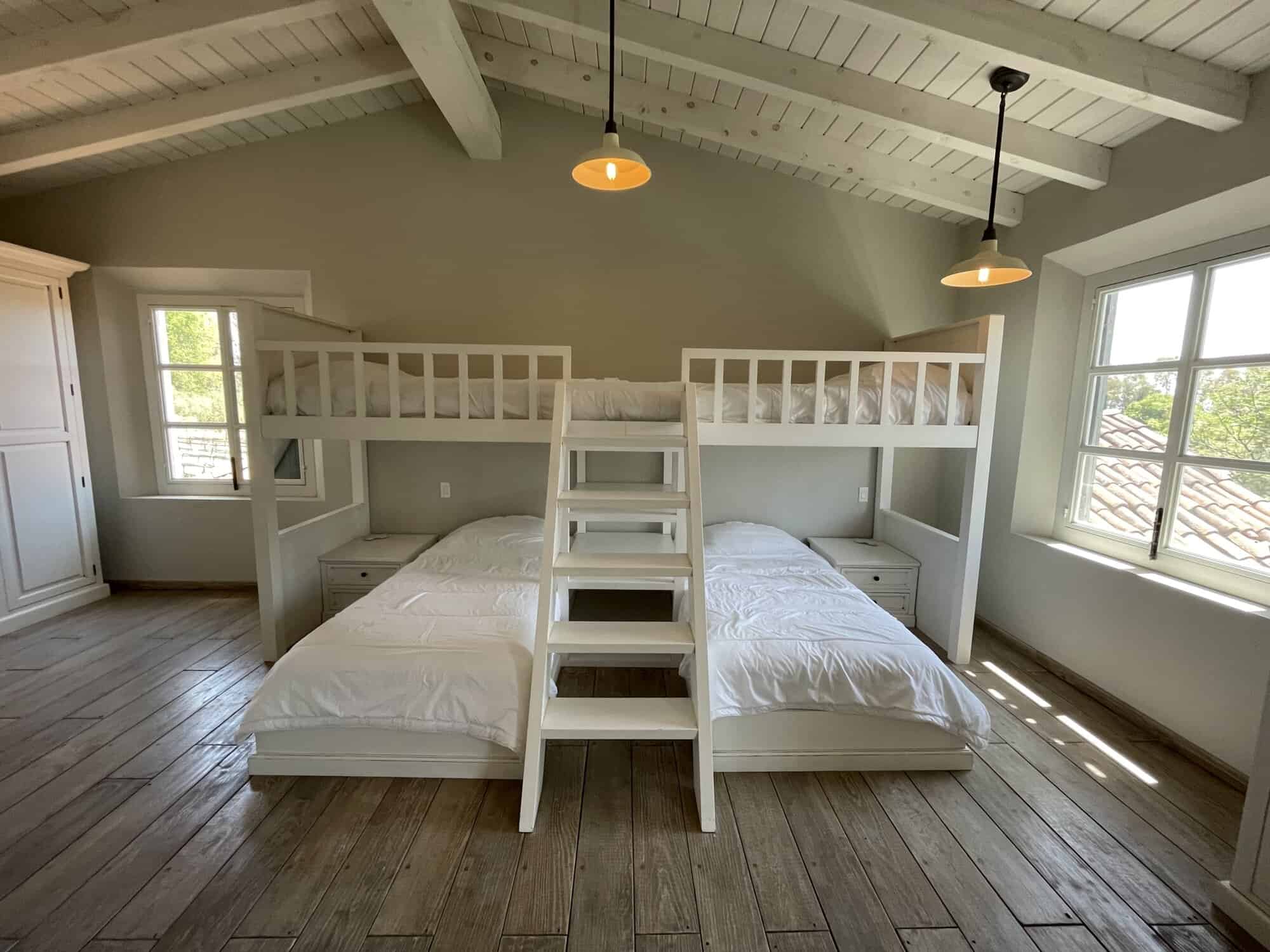 Attic bedroom with white bunk beds, large windows, and wooden flooring, offering a bright and inviti.