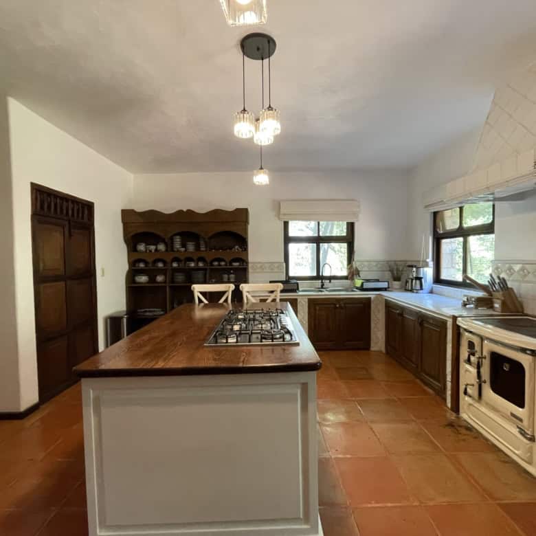 Bright kitchen with wooden cabinets, terracotta flooring, and natural light from windows.