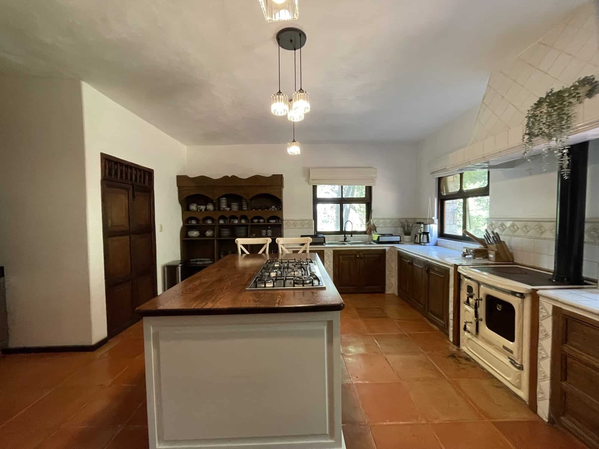 Bright kitchen with wooden cabinets, terracotta flooring, and natural light from windows.