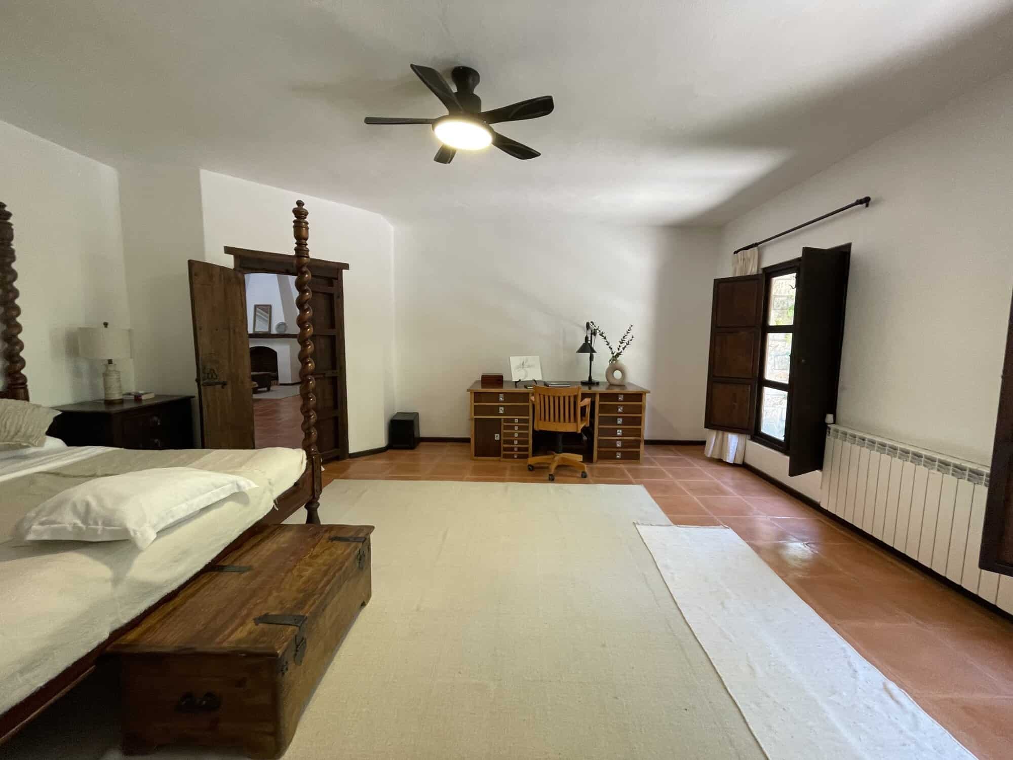Cozy bedroom featuring wooden furniture, a large window with shutters, and a ceiling fan for comfort.